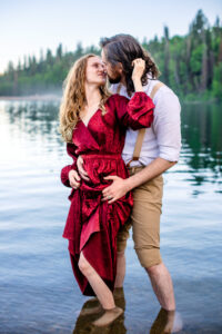 Man places hands on woman's hips while woman holds skirts of dress and places hand on his head as they both look at each other while standing in lake with trees in back, captured by engagement photographer.