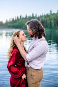 Man brushes woman's hair behind her ear as she hold skirt of dress and they both look at each other with lake and trees in back, captured by Utah engagement photographer.