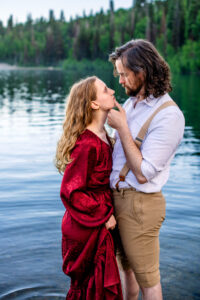 Man places hand on woman's chin as woman holds skirt of dress and they stare at each other while standing in lake, captured by Utah engagement photographer.