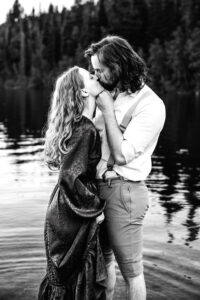 Man places hand on woman's jaw as woman holds skirt of dress and they kiss while standing in lake, captured by Utah engagement photographer in black and white.