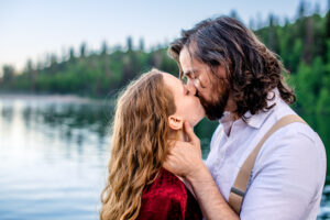 Man places hand on woman's jaw and they kiss with lake and trees in back, captured by Utah engagement photographer.