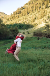 Woman in red dress runs into man's arms and man swings her with mountains and trees in the back, captured by Utah engagement photographer.