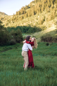 Man kisses woman's cheek and wraps arms around her as woman closes her eyes and smiles with mountains and trees in the background, captured by Utah engagement photographer.