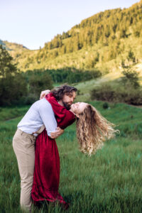 Man leans into woman in red dress and tries to kiss her as she leans back, tosses her hair, smiles, and places her arms around his neck, with trees and mountains in the back, captured by Utah engagement photographer.