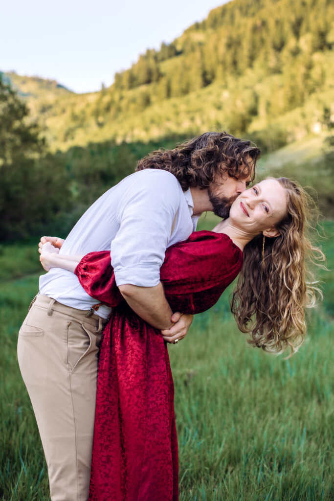 Man leans into woman in red dress and kisses her neck as she leans back, tosses her hair, smiles at camera, and places her arms around his neck, with trees and mountains in the back, captured by Utah engagement photographer.