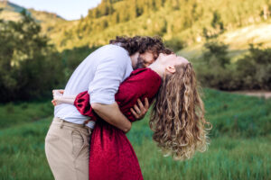 Man leans into woman in red dress and kisses her neck as she leans back, tosses her hair, smiles, and places her arms around his neck, with trees and mountains in the back, captured by Utah engagement photographer.