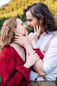 Man and woman in red dress look at each other with each of them placing a hand on the other's jaw, showcasing jewelry as woman places other hand on man's arm, captured by Utah engagement photographer.