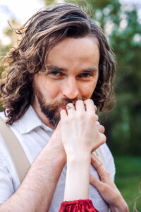 Man with curly hair and blue eyes looks and smiles at camera as he kisses woman's hand, showcasing her blue engagement ring, captured by Utah engagement photographer.