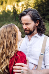 Man smiling and looking down at woman in red while he places his hands on her arms, captured by Utah engagement photographer.