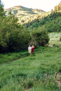 Man and woman in red dress look down and smile as they hold hands and walk along trail with tall grass and trees and mountains in back, captured by Utah engagement photographer.