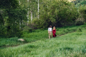 Man looks at woman in red dress as woman looks up and him an smiles as they hold hands and walk along trail with tall green grass, captured by Utah engagement photographer.