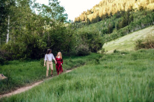 Man looks at woman in red dress as woman looks up and him an smiles while holding her dress as they hold hands and walk along trail with tall green grass, captured by Utah engagement photographer.