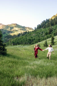 Woman in red dress holds dress and blue wildflower stem in one hand and man's hand in other hands as she leads them down a hill with man smiling at her, captured by Utah engagement photographer.