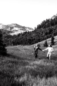 Woman in red dress holds dress and blue wildflower stem in one hand and man's hand in other hands as she leads them down a hill with man smiling at her, captured by Utah engagement photographer in black and white.