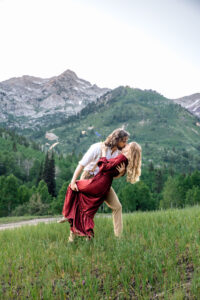 Man dips and kisses woman in red dress while holding her thigh with woman placing hand on his chest and holding blue wildflower, captured by Utah engagement photographer.