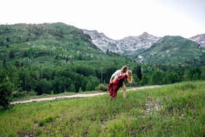 Man dips and kisses woman in red dress while holding her thigh with woman placing hand on his chest and holding blue wildflower with mountains in the back, captured by Utah engagement photographer.
