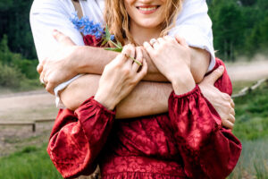 Woman in red smiling as man wraps his arms over and around her and she places hands on his arms, showing her jewelry and blue wildflower stem, captured by Utah engagement photographer.