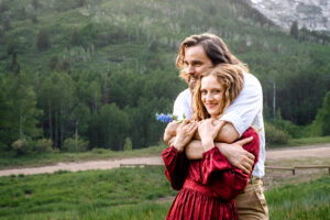 Man wraps arms over and around woman in red dress and smiles over her shoulder as woman looks and smiles at camera while holding blue wildflower stem with green hills in the back, captured by Utah engagement photographer.