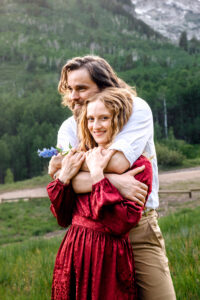 Man wraps arms over and around woman in red dress and smiles over her shoulder as woman looks and smiles at camera while holding blue wildflower stem, captured by Utah engagement photographer.