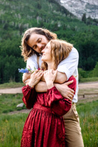 Man hugs woman from behind and wraps arms over her shoulders and kisses her cheek as woman in red dress holds his forearms and blue stemmed flower and smiles up at him, captured by Salt Lake City engagement photographer.