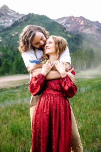 Man hugs woman from behind and wraps arms over her shoulders and kisses her cheek as woman in red dress holds his forearms and blue stemmed flower and smiles up at him with mountains in back, captured by Salt Lake City engagement photographer.
