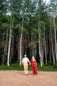 Man and woman in red dress hold hands and smile at camera as they stand on dirt road in front of tall aspen forrest, captured by Salt Lake City engagement photographer.