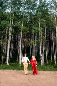 Man and woman in red dress hold hands and smile at camera as they stand on dirt road in front of tall aspen forrest, captured by Salt Lake City engagement photographer.