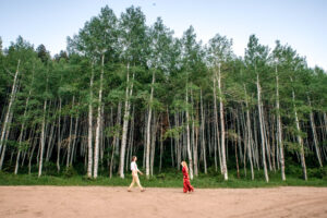 Man and woman in red dress smile and walk towards each other on dirt road in front of aspen forrest, captured by Salt Lake City engagement photographer.