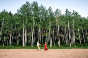 Man and woman in red dress smile and reach arms out as they walk towards each other on dirt road in front of aspen forrest, captured by Salt Lake City engagement photographer.