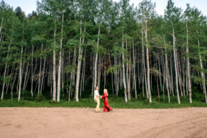 Man and woman in red dress hold hands while facing each other and smiling at each other while on dirt road in front of aspen forrest, captured by Salt Lake City engagement photographer.