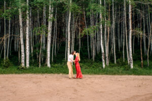 Man and woman in red dress face each other and kiss while holding hands in front of aspen trees, captured by Utah engagement photographer.