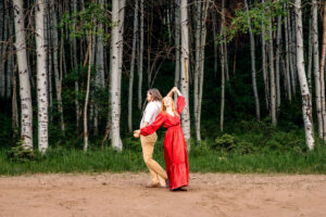 Mand and woman in red dress dance while holding hands up in air in front of aspen trees, captured by Utah engagement photographer.