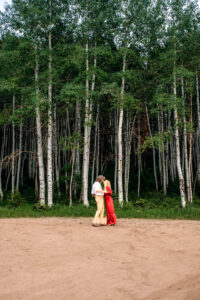 Man and woman in red dress hold hands, touch noses, and sway in front of aspen trees, captured by Utah engagement photographer.
