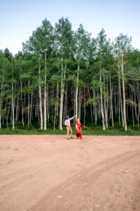 Man twirls woman in red dress on dirt path in front of aspen trees, captured by Utah engagement photographer.