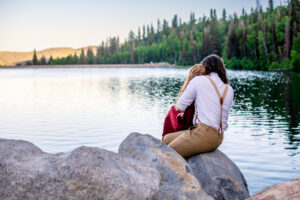Man holds woman in red dress as they sit on rock at look out at lake and trees.