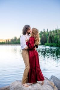 Man and woman in red dress kiss with woman placing hands on man's chest and pointing toes as the both stand on rock with lake and trees in the back, captured by Utah engagement photographer.