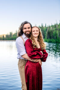 Man wraps arms around woman as they both smile at camera while standing in lake with trees in back, captured by Utah engagement photographer.
