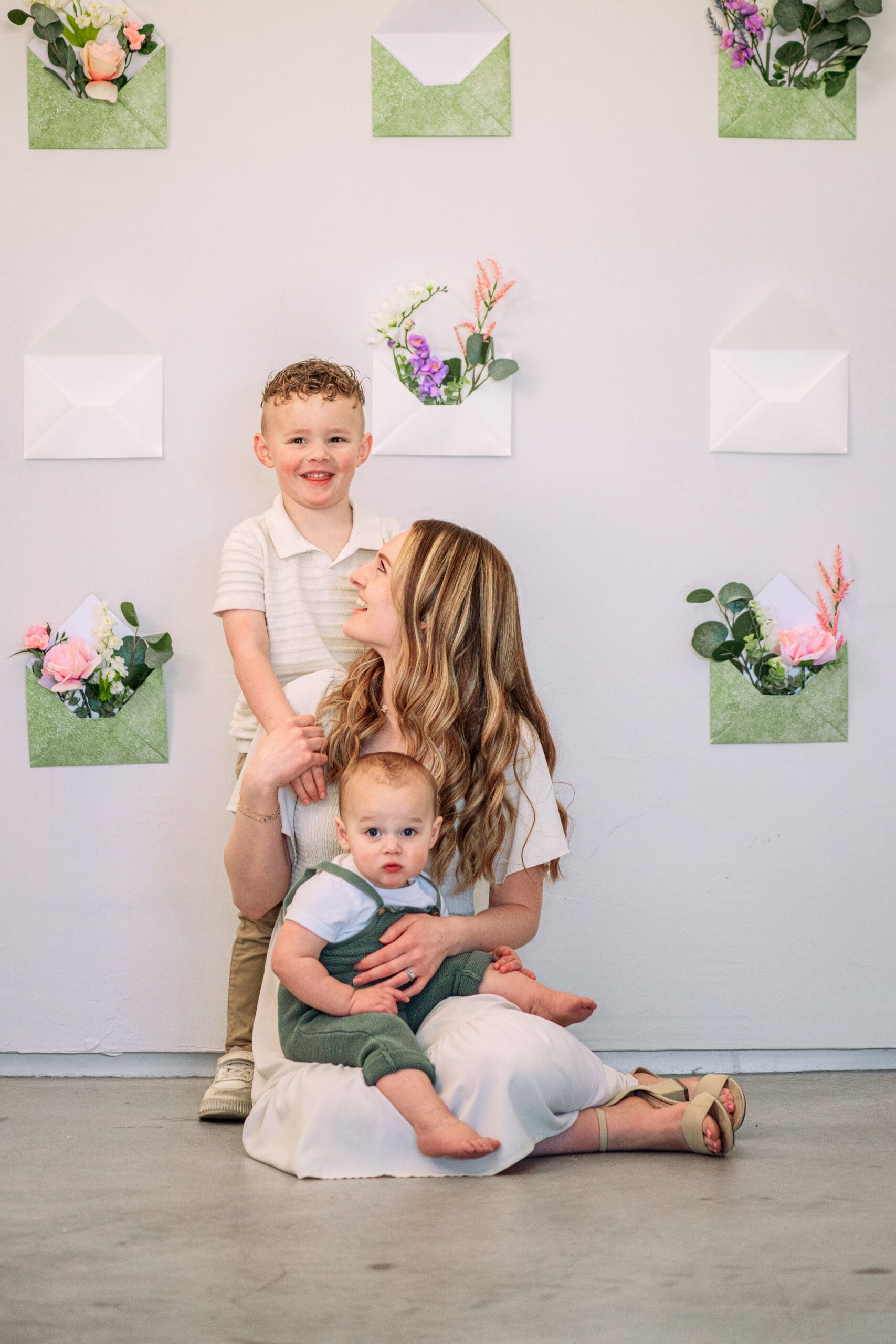 Mom sits on ground and holds young boy as she smiles up at oddler boy standing behind her in front of backdrop of cream and sage envelopes filled with flowers at a studio in Idaho Falls.