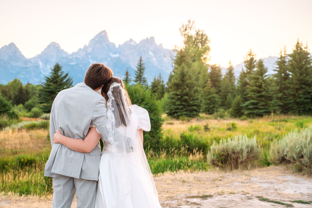 Groom and bride wraps arms around each other in side hug and look at mountains, captured by Grand Tetons wedding photographer.