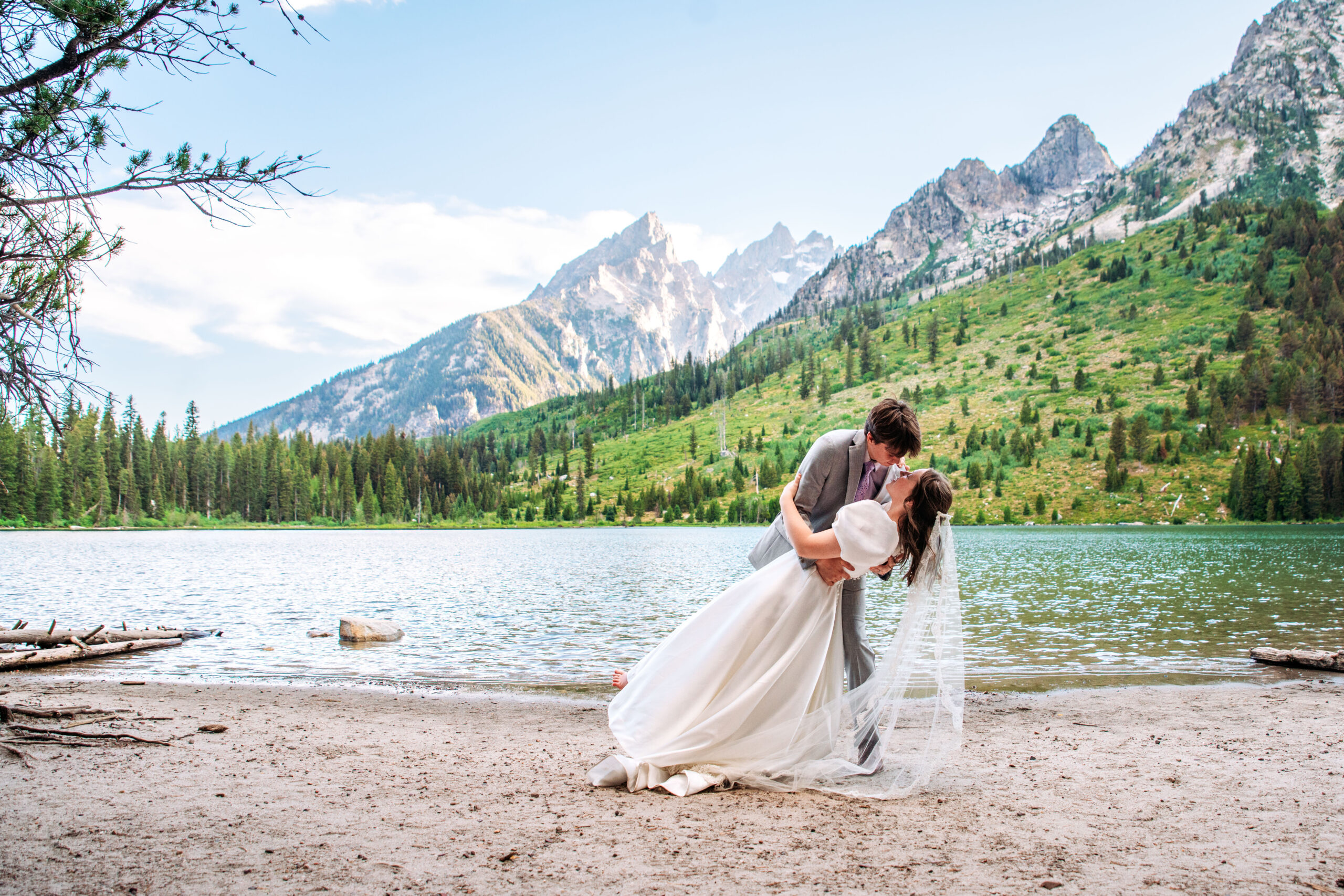 Groom dips and kisses bride on beach in front of lake and mountains, captured by Grand Tetons Wedding Photographer.