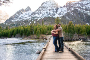Man and woman smile at camera as man wraps arms around woman from behind on bridge over river with mountains in back, captured by Grand Teton Engagement photographer.