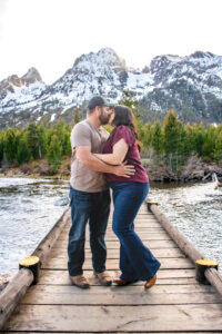 Man and woman hold each other and kiss on bridge over river with mountains in back, captured by Grand Teton Engagement photographer.