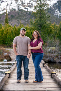 Man and woman smile at camera as woman hold man's arm and man puts hand in pocket on bridge over river with mountains in back, captured by Grand Teton Engagement photographer.