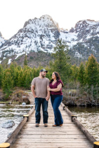 Man and woman smile at each other as woman hold man's arm and man puts hand in pocket on bridge over river with mountains in back, captured by Grand Teton Engagement photographer.