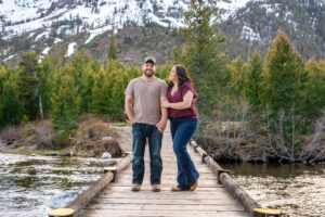 Man smiles at camera and woman smiles at man she holds his arm and man puts hand in pocket on bridge over river with mountains in back, captured by Grand Teton Engagement photographer.
