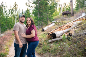 Man and woman face each other and smile at camera on trail with large moose in the background, captured by Grand Teton engagement photographer.