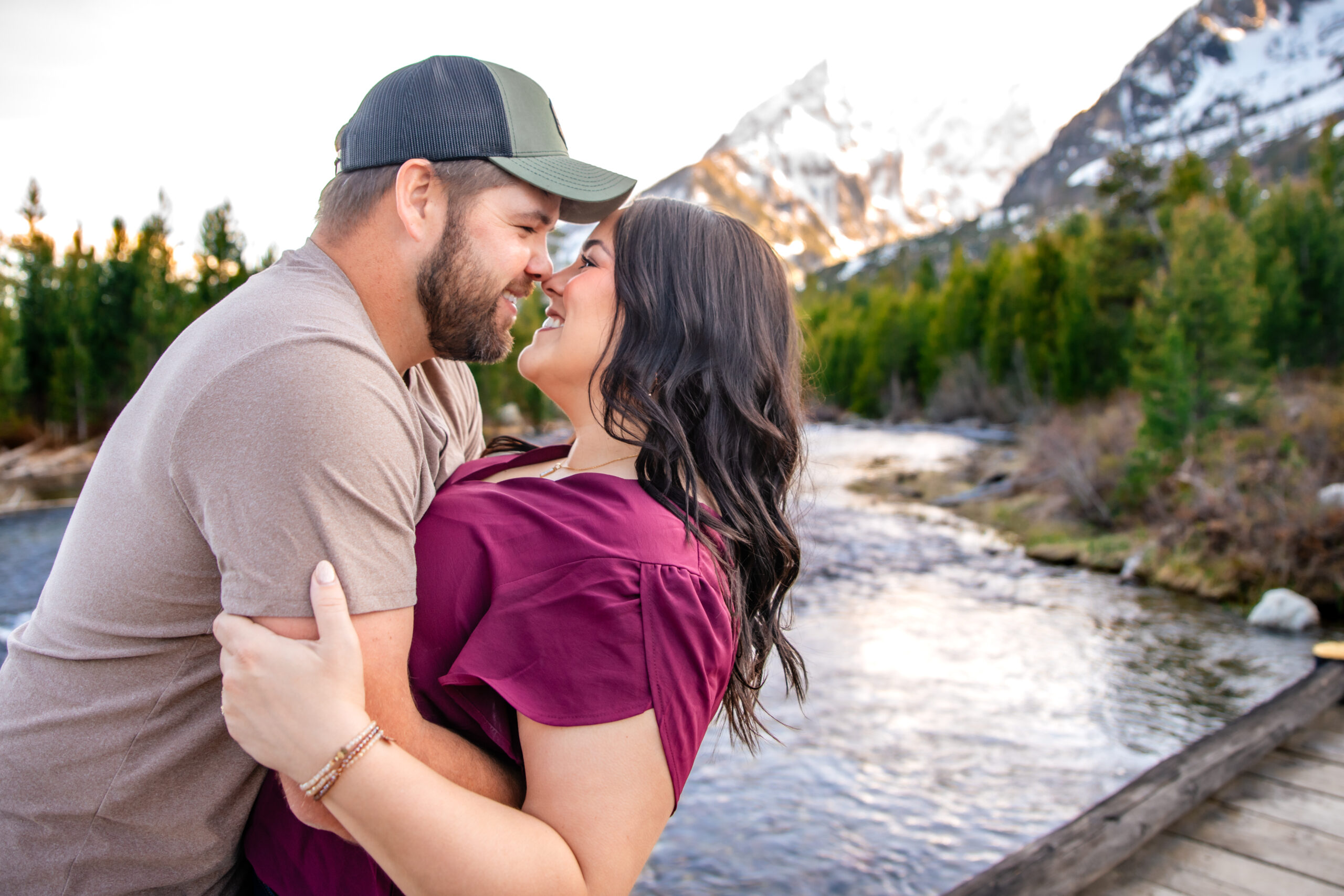 Man brings woman in and holds her as they touch noses and smile at each other on bridge over river with mountains in back, captured by Grand Teton Engagement photographer.