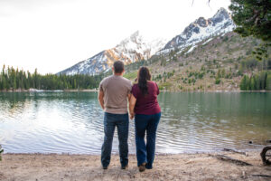 Man and woman holding hands on beach and facing lake and mountains, captured by Grand Teton engagement photographer.