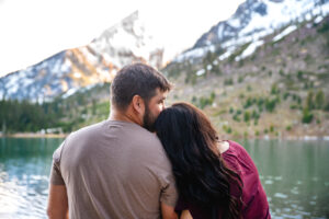 Couple facing lake and mountains as woman rests head on man's shoulder and man kisses her forehead, captured by Grand Teton engagement photographer.