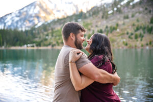 Man and woman smile and hold each other in front of lake and mountains, captured by Grand Teton engagement photographer.